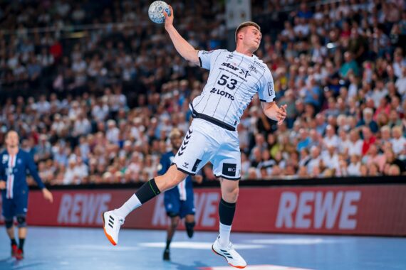 Ein Handballspieler in einem weißen Trikot springt in die Luft und bereitet sich auf den Wurf vor, während eine Menge Zuschauer von der Tribüne aus zusieht. Andere Spieler in blauen Trikots sind im Hintergrund zu sehen.