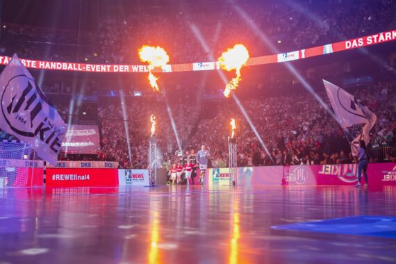 Eine vollbesetzte Arena mit jubelnden Fans, großen wehenden Fahnen und Spielern, die ein hell erleuchtetes Handballfeld betreten, während Flammen während einer energiegeladenen Veranstaltung emporschießen. Banner und digitale Anzeigen erhellen den Hintergrund.