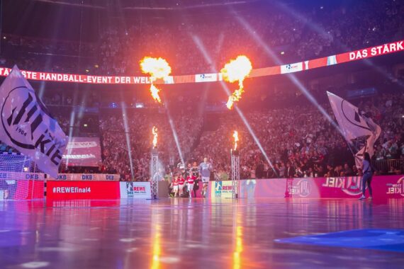 Eine hell erleuchtete Arena mit einer großen Menschenmenge, brennenden Fackeln und zwei wehenden Fahnen, während eine Handballmannschaft das Spielfeld für ein Turnier betritt. Die Atmosphäre ist lebhaft und festlich.