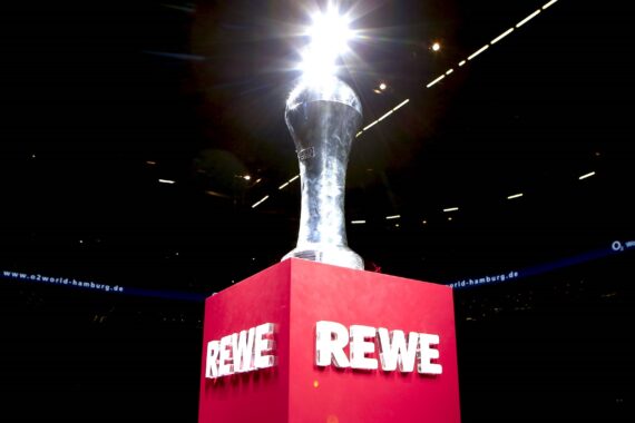 Auf einem roten Sockel steht eine silbern glänzende Trophäe mit dem Schriftzug REWE auf allen sichtbaren Seiten, die von einem hellen Scheinwerfer vor einem dunklen Hintergrund angestrahlt wird.