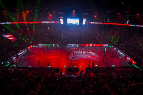 Eine große, mit Zuschauern gefüllte Halle beobachtet ein hell erleuchtetes Handballfeld, auf dem zwei Mannschaften und Offizielle vor einem Spiel eine Zeremonie abhalten. Rote und grüne Lichter und Bildschirme zeigen Informationen über die Veranstaltung in der Halle an.