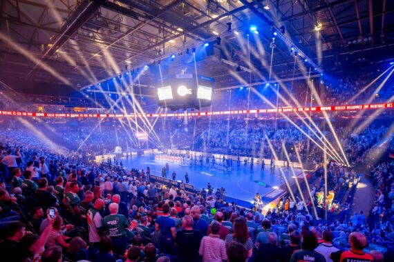 Ein großes, volles Hallenstadion mit hellen Scheinwerfern und bunten Lichtern, die ein Handballfeld beleuchten, während die Zuschauer zusehen und die Spieler sich vor dem Spiel aufstellen. Die Atmosphäre ist energiegeladen und festlich.