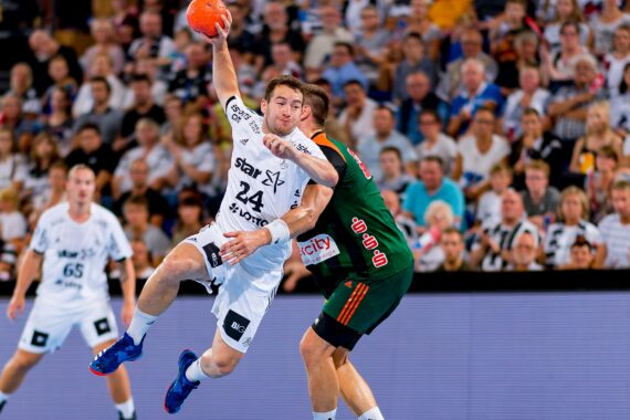 Ein Handballspieler in einem weißen Trikot springt, um den Ball zu werfen, während er von einem Spieler in einem dunkelgrünen Trikot eng verteidigt wird, während die Zuschauer im Hintergrund zusehen.
