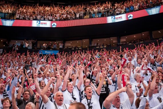 Eine große Menge begeisterter, meist weiß gekleideter Fans jubelt mit erhobenen Armen und Klatschstöcken in einem überfüllten Hallenstadion und sorgt für eine lebhafte und energiegeladene Atmosphäre.