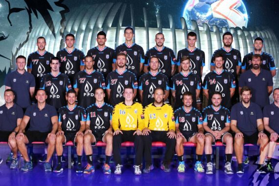 Eine Handballmannschaft posiert für ein Gruppenfoto in schwarz-gelben Trikots, die in drei Reihen angeordnet sind, mit einem futuristischen Stadion und einer Löwengrafik im Hintergrund.