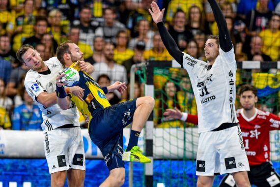 Ein Handballspieler in einem blau-gelben Trikot versucht zu schießen, während er von zwei Spielern in weißen Trikots verteidigt wird, während im Hintergrund ein Publikum und ein Torwart zusehen.