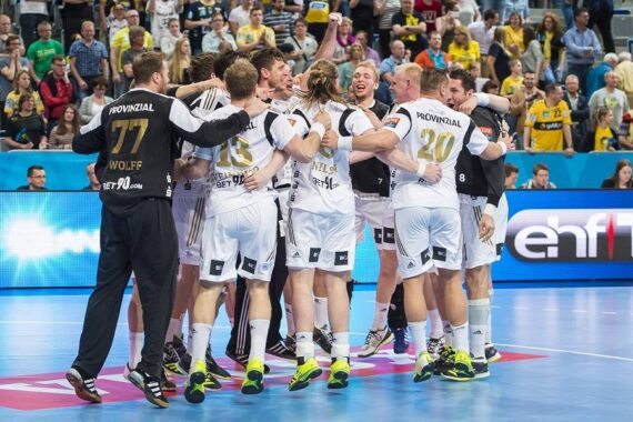 Eine Gruppe männlicher Handballspieler in weißen Trikots feiert auf einem Hallenplatz, lächelt und umarmt sich, während die Fans im Hintergrund von der Tribüne aus zusehen.