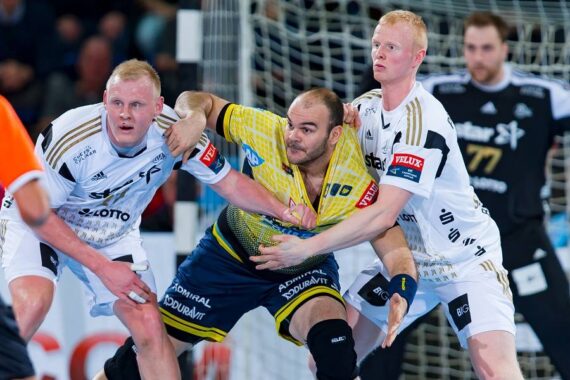 Drei männliche Handballspieler in Aktion während eines Spiels. Zwei Spieler in weißen Trikots verteidigen gegen einen Spieler in einem gelben Trikot, der den Ball hält und versucht, durchzubrechen. Die Zuschauer sind im Hintergrund verschwommen.