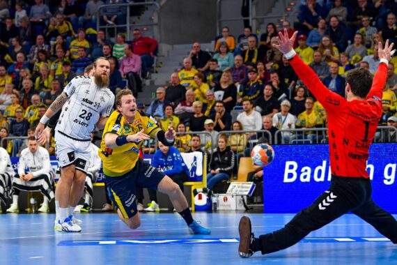 Ein Handballspieler in einem gelben Trikot springt ab und schießt in Richtung Tor, während ein Torwart in einem roten Trikot seine Arme ausbreitet, um zu blocken. Ein anderer Spieler in einem weißen Trikot steht in der Nähe. Die Zuschauer füllen die Plätze in der Arena im Hintergrund.