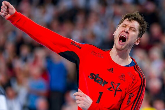 Ein männlicher Handballspieler in einem leuchtend roten Trikot mit Stern und der Nummer 1 jubelt mit geballter Faust und geschlossenen Augen und scheint während eines Spiels vor Begeisterung zu schreien. Im Hintergrund ist eine unscharfe Menschenmenge zu sehen.