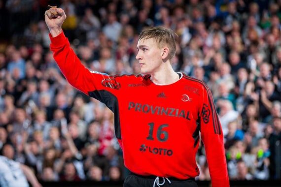 Ein Handballspieler in einem roten Trikot mit der Nummer 16 und der Aufschrift PROVINZIAL reckt die Faust in die Luft. Er steht auf dem Spielfeld, während die Zuschauermenge im Hintergrund verschwommen ist.