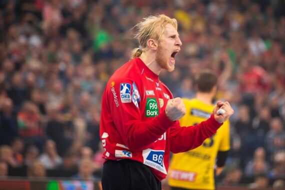 Ein männlicher Handballspieler in einem roten Trikot jubelt leidenschaftlich mit geballten Fäusten und offenem Mund, wahrscheinlich schreiend. Im Hintergrund sind eine verschwommene Menschenmenge und ein weiterer Spieler in einem gelben Trikot zu sehen.