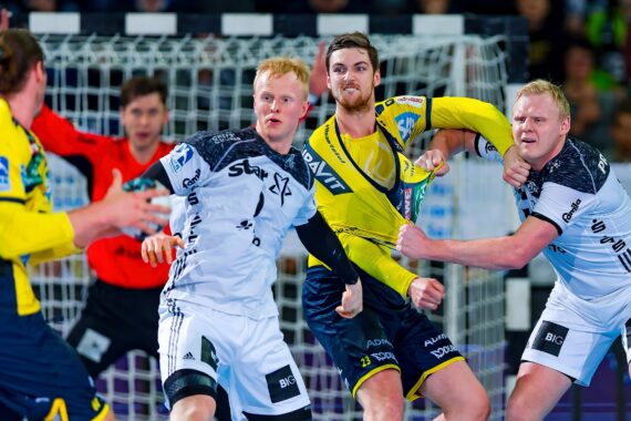 Drei männliche Handballspieler in Aktion in der Nähe des Tores, zwei in weißen Trikots verteidigen gegen einen Spieler in einem gelben Trikot, während ein Schiedsrichter und ein Torwart im Hintergrund zusehen.