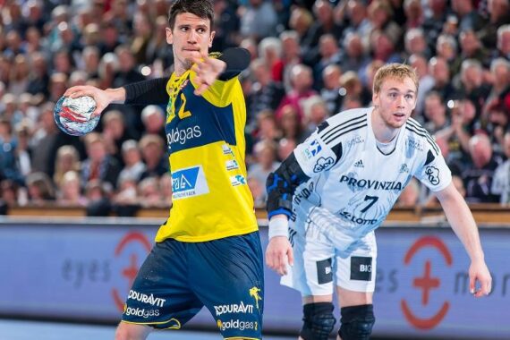 Ein Handballspieler in einem gelb-blauen Trikot bereitet sich auf den Wurf vor, während ein Verteidiger in einem weiß-schwarzen Trikot aufmerksam zusieht. Im Hintergrund ist eine Menge von Zuschauern zu sehen.