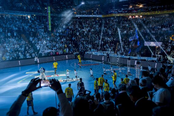 Eine überfüllte Halle mit hellen Lichtern und Konfetti, in der sich Sportler in gelben und weißen Uniformen auf einem glänzenden Spielfeld aufstellen, während ein kostümiertes Maskottchen daneben steht. Die Fans füllen die Tribünen, einige halten den Moment mit ihren Handys fest.