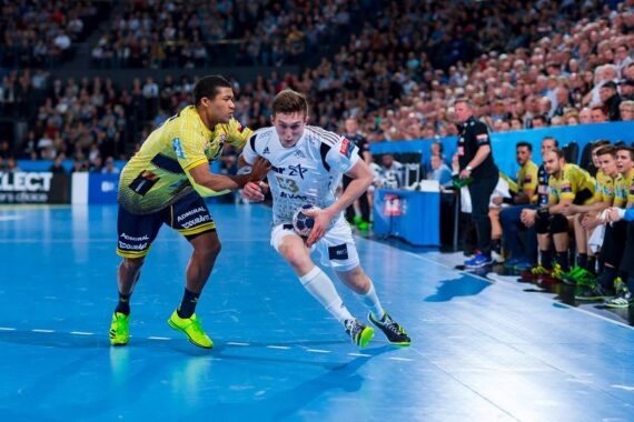 Zwei männliche Handballspieler kämpfen auf einem blauen Spielfeld um den Ball; der eine in einem gelben Trikot versucht, den anderen in einem weißen Trikot zu blockieren, während eine große Menschenmenge von der Tribüne aus zusieht.