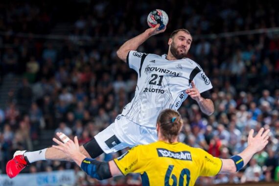 Ein Handballspieler in einem weißen Trikot springt in der Luft und bereitet den Wurf vor, während ein Spieler in einem gelben Trikot den Ball mit erhobenen Armen abwehrt. Im Hintergrund ist eine Menge von Zuschauern zu sehen.
