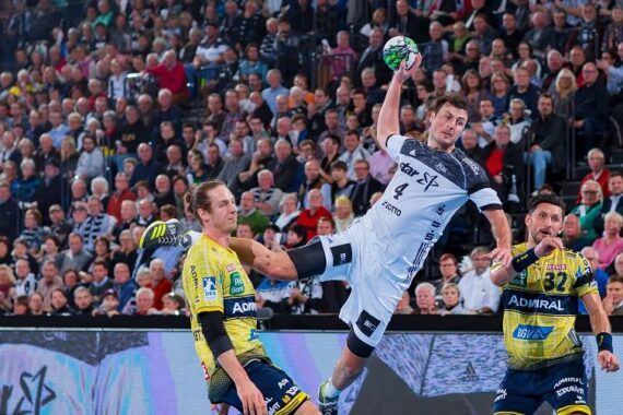 Ein Handballspieler in einem weißen Trikot springt in die Luft, um zu schießen, während zwei Verteidiger in gelben Trikots versuchen, ihn abzuwehren; im Hintergrund schaut eine große Menschenmenge aufmerksam zu.