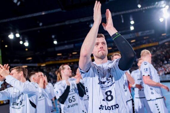 Eine Gruppe männlicher Handballspieler in weißen Trikots klatscht und jubelt auf einem Hallenplatz, wobei im Hintergrund eine große Menschenmenge und helle Lichter zu sehen sind. Der vorderste Spieler hebt die Hände über den Kopf und applaudiert.