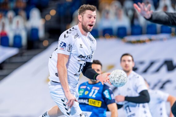Ein männlicher Handballspieler in einem weißen Trikot bereitet sich darauf vor, den Ball während eines Spiels zu passen oder zu schießen, während im Hintergrund Mannschaftskameraden und ein Gegner zu sehen sind.
