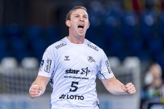 Ein männlicher Handballspieler in einem weißen Trikot mit der Nummer 5 jubelt mit geballten Fäusten und offenem Mund und scheint zu schreien. Der Hintergrund ist unscharf, es sind blaue Sitze und undeutliche Figuren zu sehen.