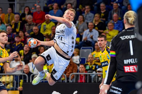 Ein Handballspieler in einem weißen Trikot springt, um den Ball zu schießen, während zwei Verteidiger in Gelb und ein Torwart in Schwarz während eines Spiels in einer überfüllten Halle aufmerksam zusehen.