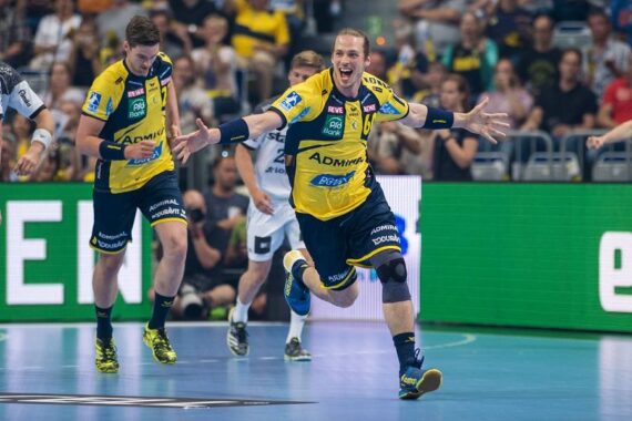Ein männlicher Handballspieler in einem gelb-blauen Trikot jubelt mit ausgestreckten Armen auf einem Hallenplatz, während Mitspieler und Gegner zusehen. Im Hintergrund ist das Publikum zu sehen.