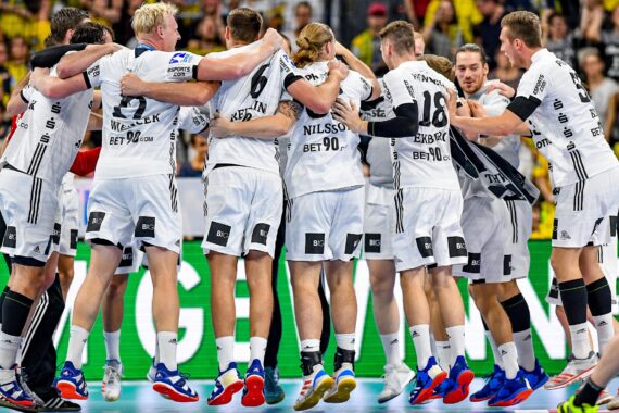Eine Gruppe männlicher Handballspieler in weißen Trikots steht in einer Halle und bereitet sich auf ein Spiel vor, während im Hintergrund eine Menschenmenge zusieht.