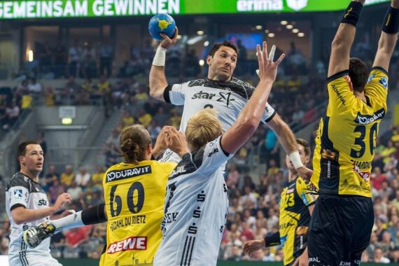 Ein Handballspieler in einem weißen Trikot springt, um den Ball zu werfen, während drei Spieler in gelben Trikots versuchen, ihn zu blockieren. Die Aktion findet in einer überfüllten Halle statt.