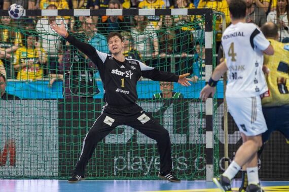 Ein Handballtorwart in schwarzer Uniform bereitet sich darauf vor, einen Schuss mit weit ausgestreckten Armen vor dem Tor abzublocken, während sich ein gegnerischer Spieler in Weiß mit dem Ball nähert. Die Zuschauer schauen von der Tribüne im Hintergrund zu.