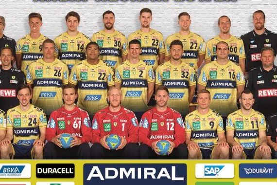 Eine Handballmannschaft der Männer in gelben Trikots posiert für ein offizielles Gruppenfoto mit Trainern und Mitarbeitern. Drei Spieler in roten Trikots halten blaue Handbälle. Am unteren Rand sind Sponsorenlogos zu sehen. Die Gruppe steht vor einem weißen Hintergrund.