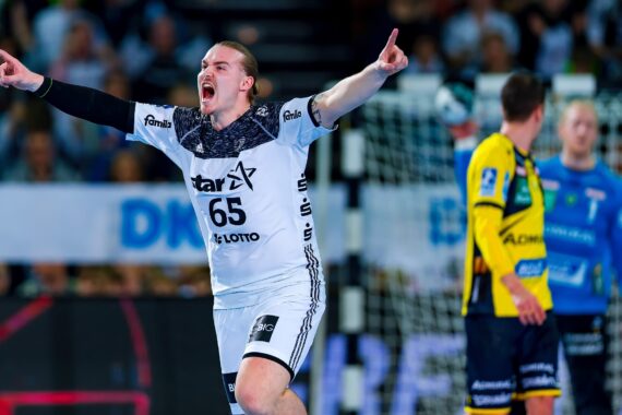 Ein männlicher Handballspieler in einem weißen Trikot mit der Nummer 65 feiert mit erhobenen Armen, während andere Spieler in gelben und blauen Trikots im Hintergrund auf dem Spielfeld stehen.