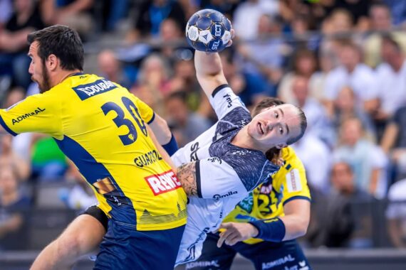 Ein Handballspieler in einem weiß-blauen Trikot springt und bereitet sich auf den Wurf vor, während er während eines Spiels von einem Spieler in einem gelb-blauen Trikot eng verteidigt wird, mit einer unscharfen Zuschauermenge im Hintergrund.