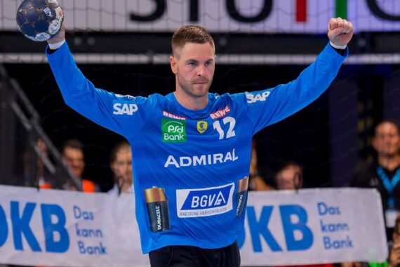 Ein Handballtorwart in einem blauen Trikot mit der Nummer 12 hebt während eines Spiels den Arm und hält einen Ball. Im Hintergrund sind Werbebanner und Zuschauer zu sehen.