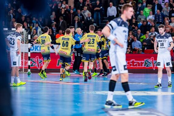 Eine Gruppe von Handballspielern in gelben und blauen Trikots feiert auf dem Spielfeld, während Spieler in weißen Trikots im Vordergrund stehen; im Hintergrund sieht eine Menschenmenge zu.