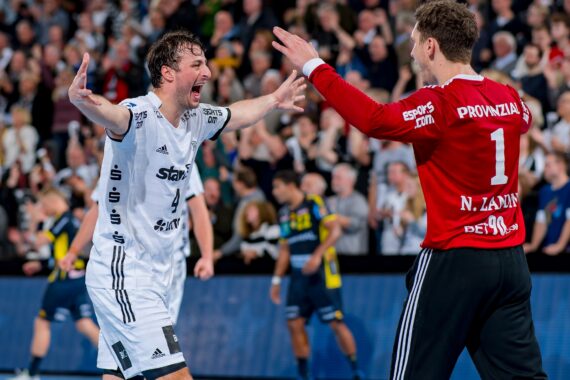 Zwei männliche Handballspieler in Uniformen feiern ausgelassen auf dem Spielfeld, wobei sie sich mit weit geöffneten Armen gegenüberstehen. Eine jubelnde Menge und andere Spieler sind im unscharfen Hintergrund zu sehen.