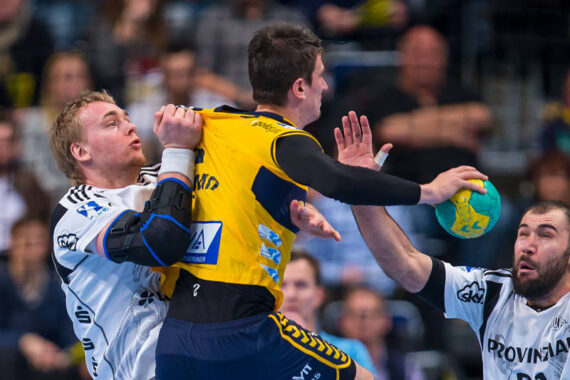 Zwei Handballspieler in weißen Trikots verteidigen sich gegen einen Spieler in einem gelben Trikot, der springt, einen grün-gelben Ball in der Hand hält und zu schießen versucht. Die Szene ist intensiv und actionreich, mit unscharfen Zuschauern im Hintergrund.
