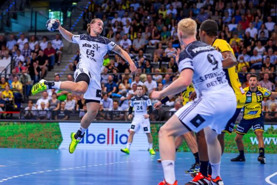 Ein Handballspieler in einem weißen Trikot springt und wirft den Ball in Richtung Tor, während Verteidiger und Mannschaftskameraden zusehen. Das Spiel findet in einer überfüllten Halle mit blauem Spielfeld statt.