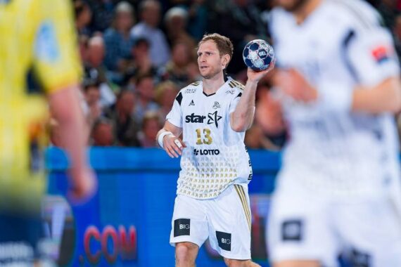 Ein männlicher Handballspieler in einem weißen Trikot bereitet sich darauf vor, den Ball während eines Spiels zu werfen. Im Hintergrund sind andere Spieler und eine Menge von Zuschauern zu sehen.