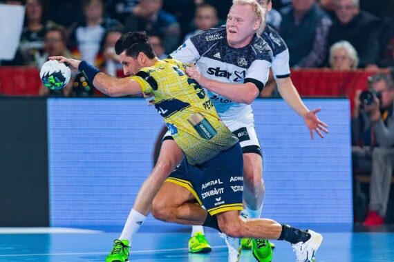 Zwei männliche Handballspieler liefern sich einen intensiven Wettkampf. Der eine in einem gelben Trikot stürzt sich mit dem Ball nach vorne, während ein Spieler in einem weiß-schwarzen Trikot versucht, ihn während eines Spiels zu blocken.