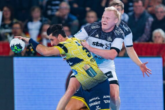 Ein Handballspieler in einem gelb-blauen Trikot versucht einen Schuss, während er von einem gegnerischen Spieler in einem weiß-schwarzen Trikot während eines Spiels eng verteidigt wird, wobei im Hintergrund Zuschauer zu sehen sind.
