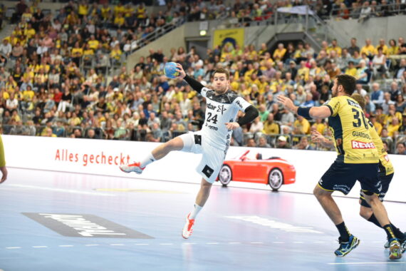 Ein Handballspieler in einem weißen Trikot springt, um den Ball zu werfen, während ein Verteidiger in einem dunklen Trikot versucht, ihn zu blockieren. Das Spiel findet in einer mit Zuschauern gefüllten Halle statt.