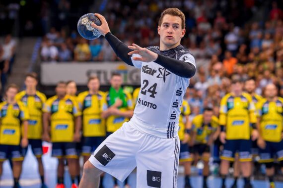 Ein Handballspieler in einem weißen Trikot bereitet sich darauf vor, den Ball während eines Spiels zu werfen, während eine Gruppe von Spielern in gelben und blauen Trikots und eine jubelnde Menge im Hintergrund zu sehen sind.