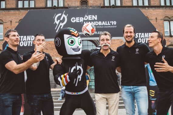 Fünf Männer stehen lächelnd beieinander und haben einen Arm um eine Person in einem Roboter-Maskottchen-Kostüm gelegt. Sie stehen draußen, vor einem Gebäude und einem Banner für die Handball-Weltmeisterschaft 2019 in Dänemark.