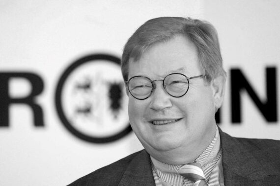 Ein Mann mittleren Alters mit Brille und Anzug lächelt freundlich. Im Hintergrund sind große, teilweise sichtbare schwarze Buchstaben auf einer weißen Wand zu sehen. Das Bild ist in schwarz-weiß gehalten.