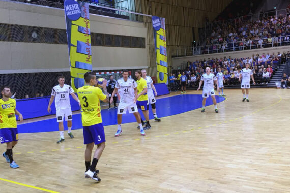Ein Handballspiel der Männer in einer Sporthalle; ein Spieler in einem gelben Trikot bereitet sich auf den Wurf vor, während Spieler in weißen Trikots verteidigen. Die Zuschauer füllen die Tribünen im Hintergrund. An den Wänden hängen leuchtende Fahnen.