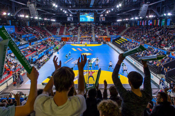 Eine überfüllte Halle mit jubelnden und mit Schaumstoffstöcken wedelnden Fans, die ein Handballspiel auf einem blauen Spielfeld unter heller Beleuchtung verfolgen. Die Tribünen sind mit Zuschauern gefüllt.