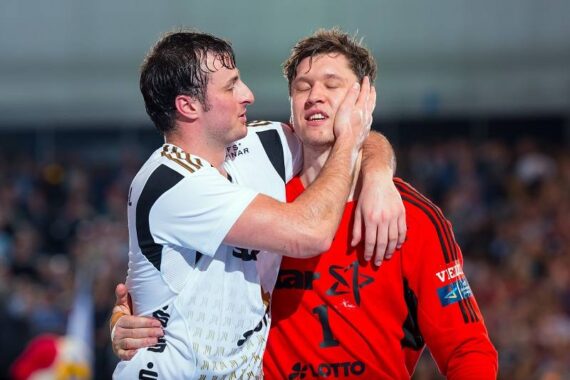 Zwei männliche Handballspieler, einer in einem weißen Trikot und der andere in einem roten Torwarttrikot, umarmen sich auf dem Spielfeld. Der Spieler in Weiß hält sanft das Gesicht des anderen, während sie beide emotional wirken.