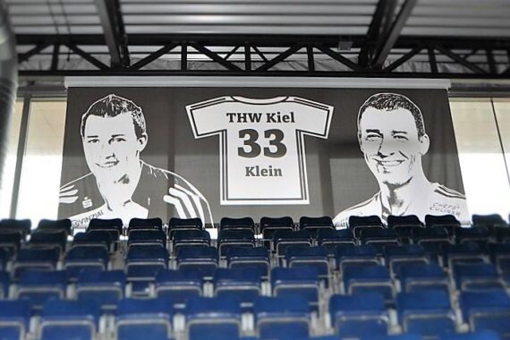 Auf einem Banner über den leeren Sitzen des Stadions ist ein Trikot mit der Aufschrift THW Kiel 33 Klein zwischen schwarz-weißen Zeichnungen von zwei lächelnden Männern zu sehen.