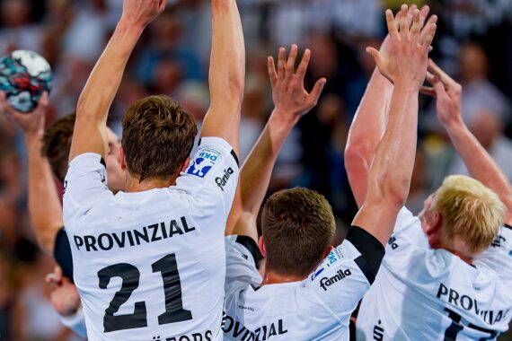 Eine Gruppe männlicher Sportler in weißen Trikots mit der Aufschrift PROVINZIAL auf dem Rücken springt mit erhobenen Armen und versucht, einen Schuss während eines Handballspiels abzuwehren.